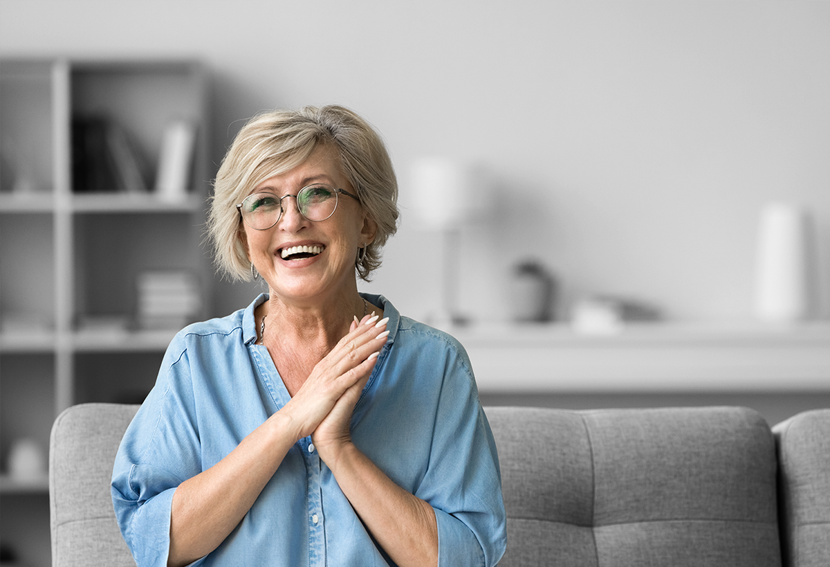 Smiling older woman with short blonde hair and glasses sits on a sofa with hands clasped.