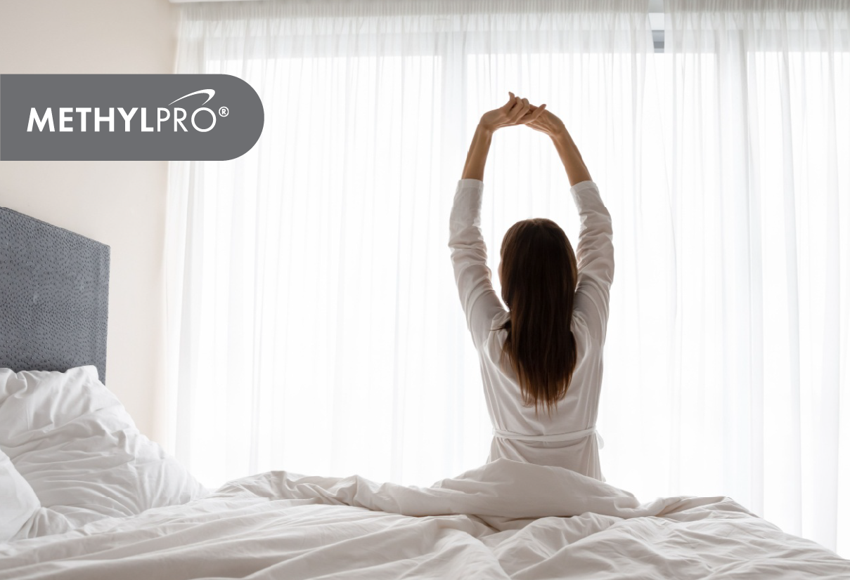Person stretches arms above head on a bed in a bright sunlit bedroom with a MethylPro logo overlay