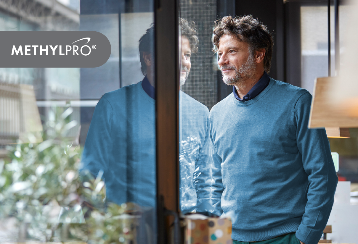 A man in a blue sweater stands near a glass storefront, looking out with a thoughtful smile; MethylPro branding is visible.