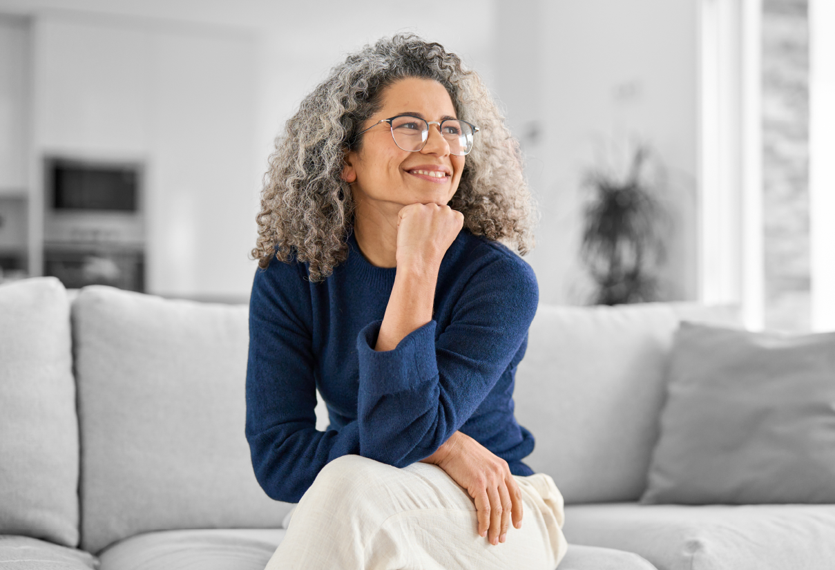 Smiling woman with curly gray hair and glasses sits on a light sofa in a bright living room.