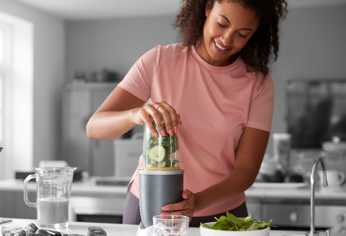 Woman in a pink shirt blends a green smoothie in a blender on the kitchen counter.