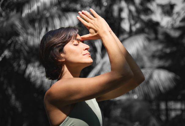 Woman meditates outdoors with arms raised among tropical plants.
