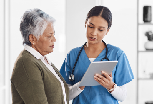 Healthcare professional in blue scrubs discusses with an elderly patient while showing a tablet.