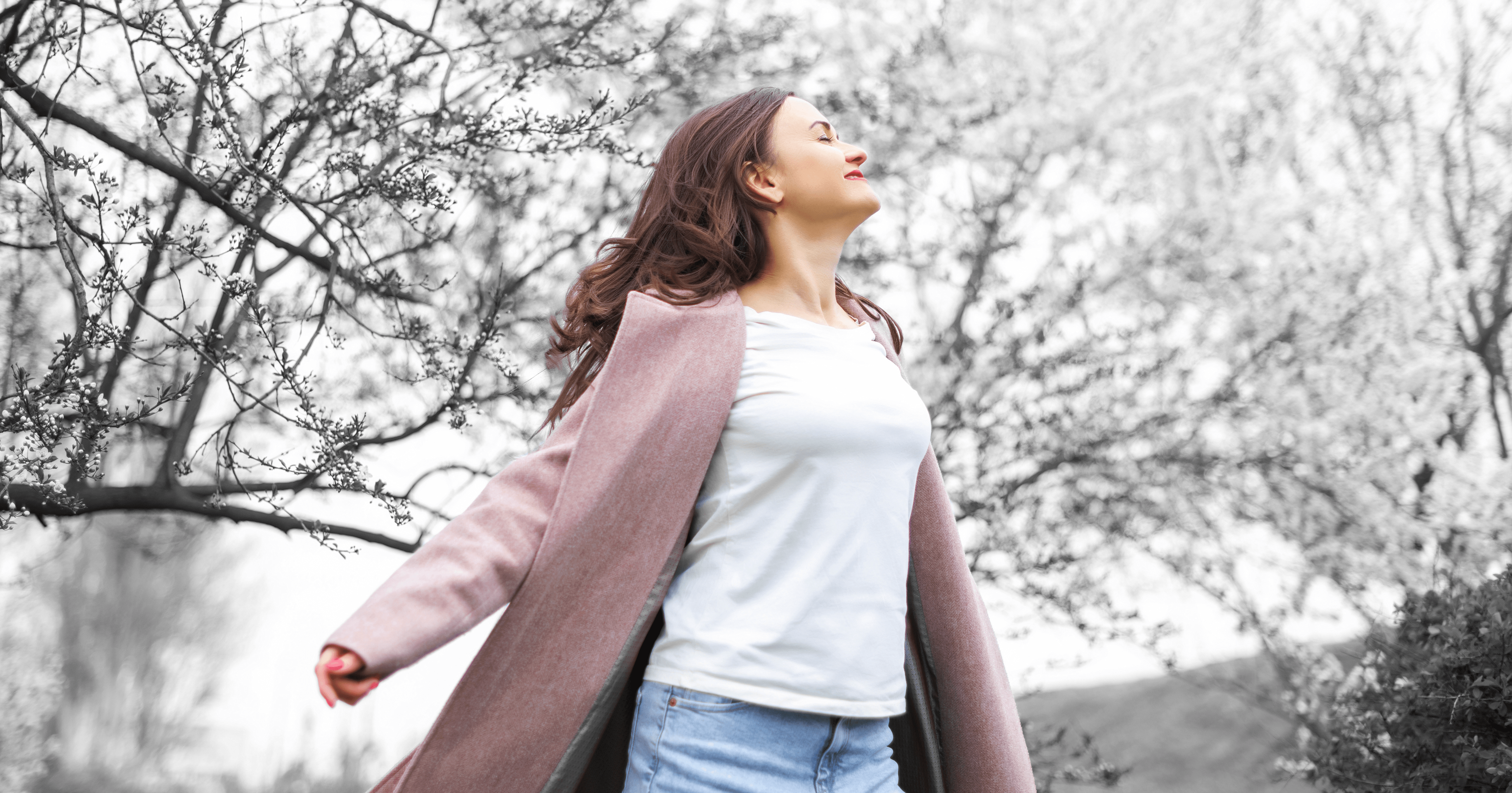 Woman in a pink coat and white shirt stands outdoors with arms outstretched among blossoming trees.