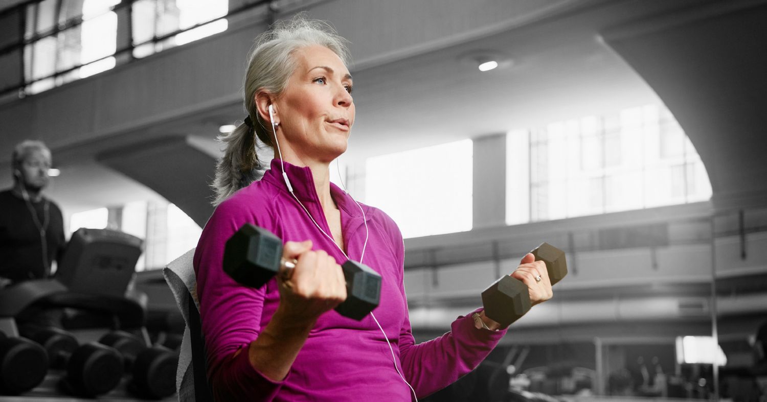 Older woman in a magenta athletic top performing bicep curls with dumbbells in a gym while wearing earbuds