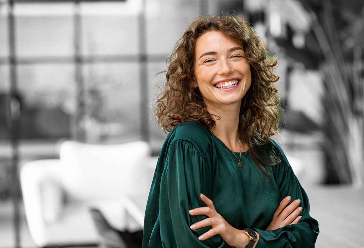 Portrait of a smiling woman with curly hair wearing a green blouse indoors with arms crossed