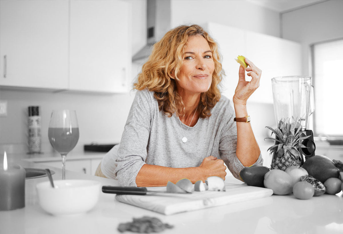 Smiling woman with curly blond hair in a teal top leans on a kitchen counter holding a sticky note.