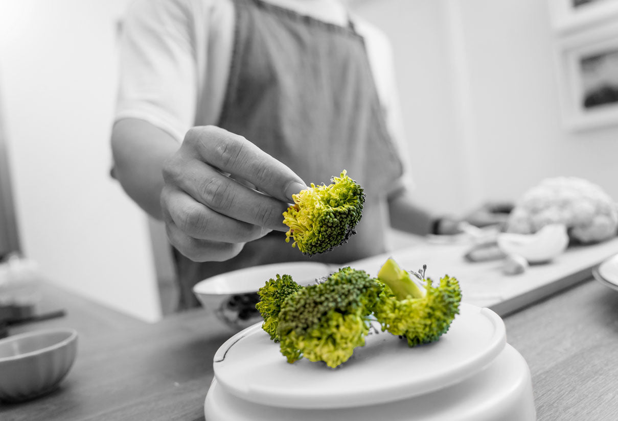 Person in apron places a bright green broccoli floret onto a plate in the kitchen.
