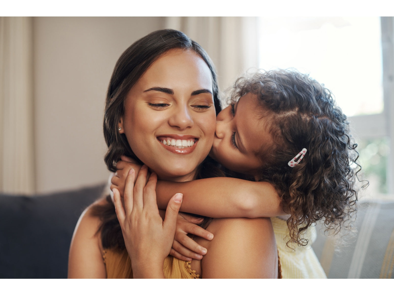 Smiling mother receives a kiss on the cheek from her young daughter who wraps her arms around her neck.