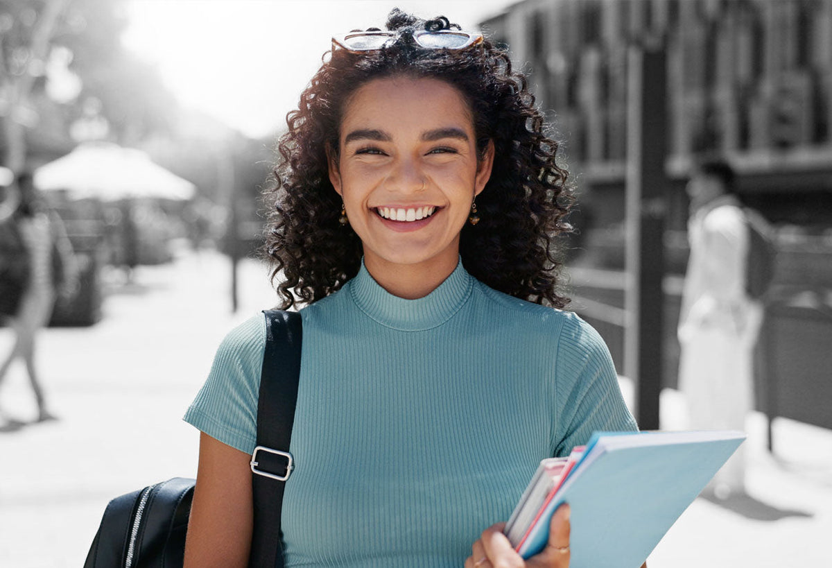 Smiling woman with curly dark hair in a teal turtleneck walks on a city sidewalk carrying books and a backpack.