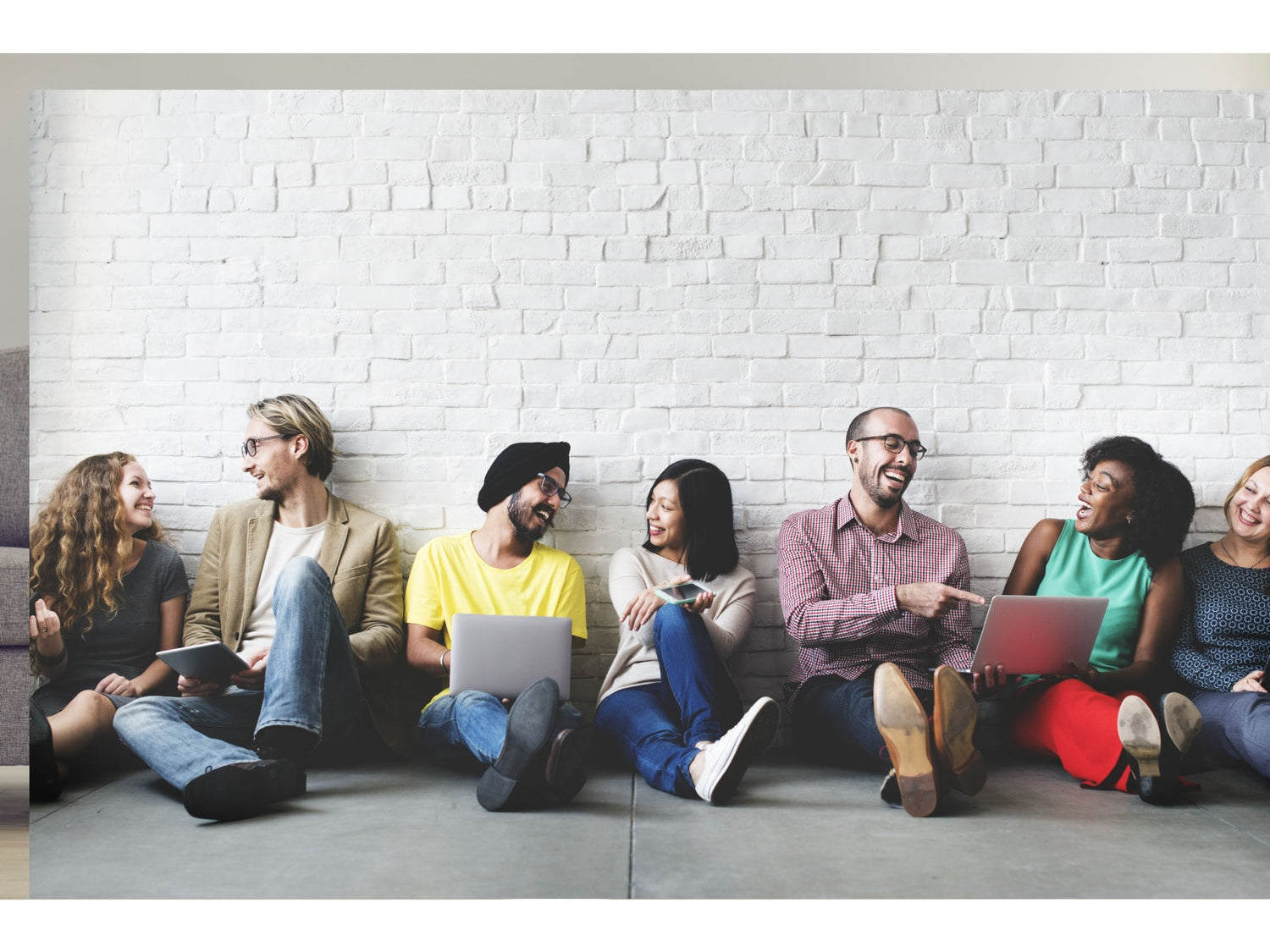 Diverse group of friends sitting against a white brick wall, smiling and chatting with laptops and tablets.