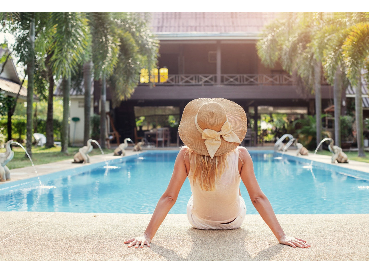 Woman sits at the edge of a resort pool wearing a wide straw hat with a bow.