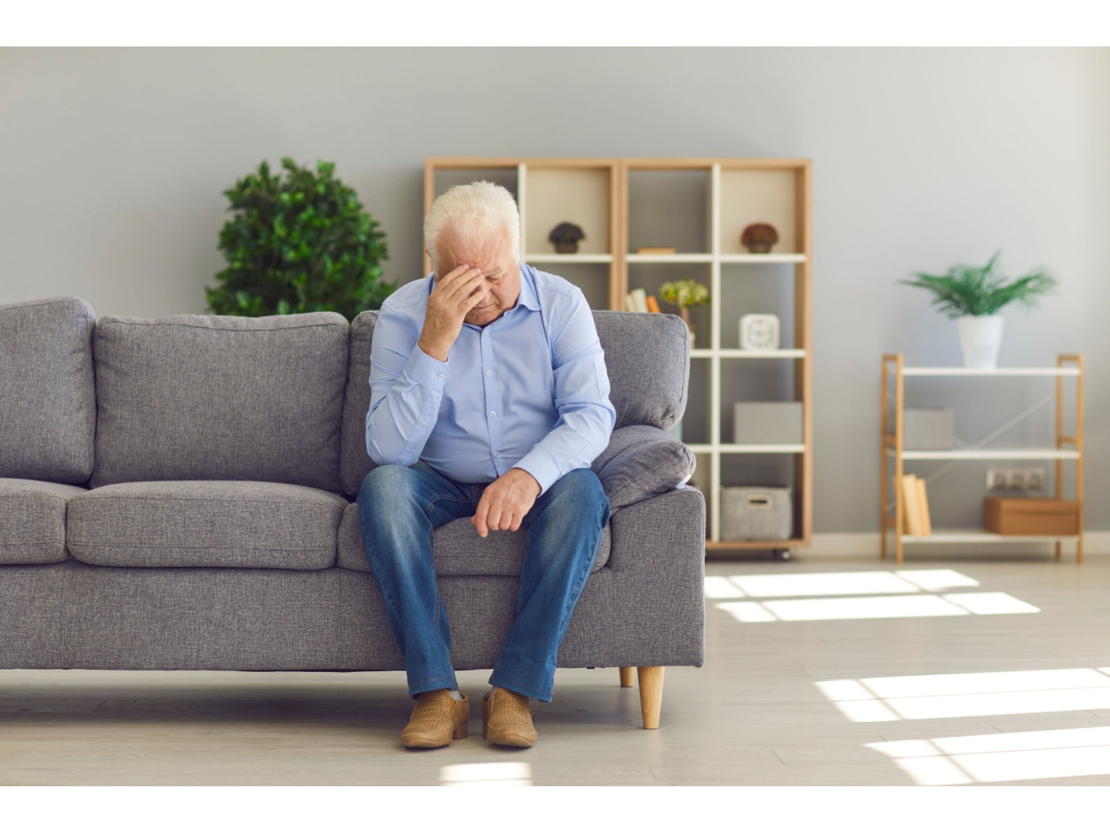 An older man sits on a gray sofa with one hand on his forehead, looking distressed in a bright living room.