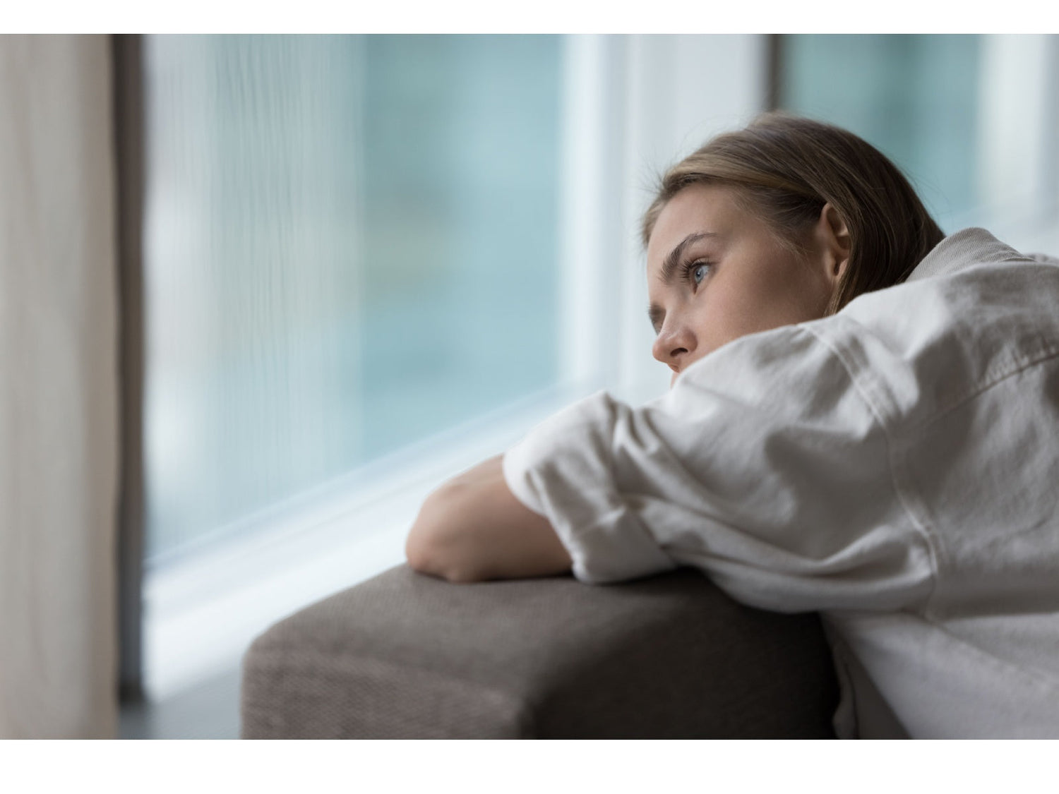 Woman resting on a sofa by a window, gazing outward with a thoughtful somber expression.