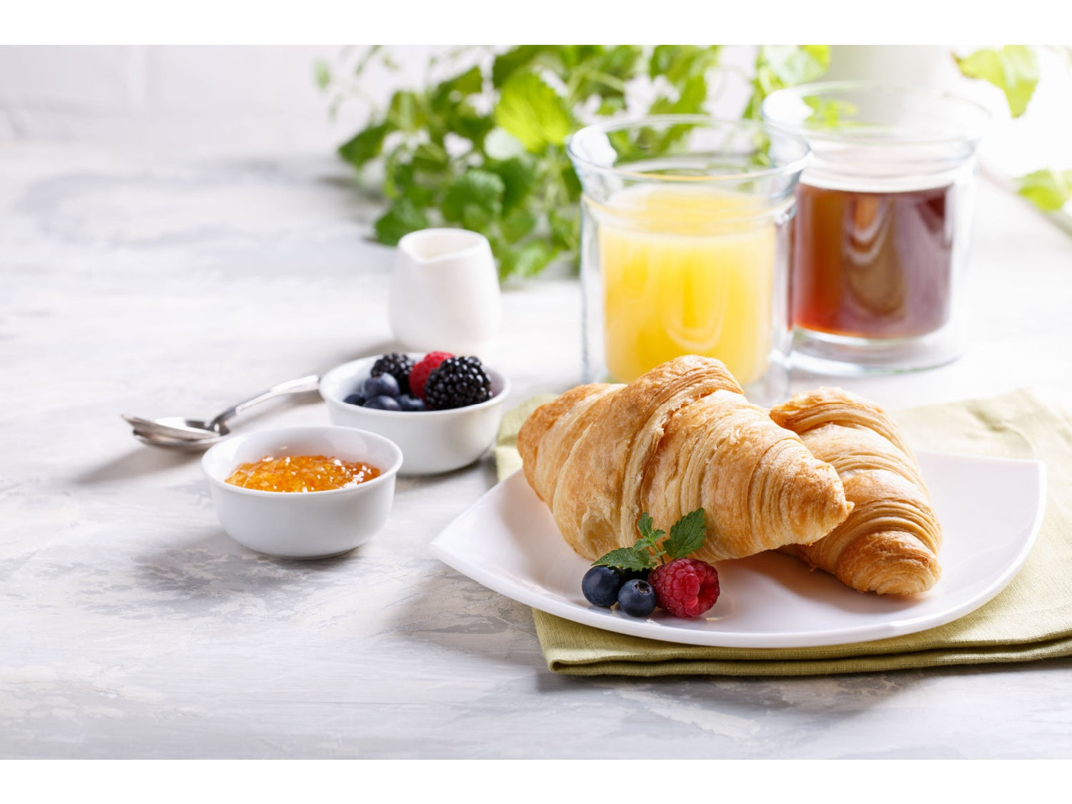 Golden croissant on a white plate set on a pale green napkin, with blueberries, raspberries, jam, and two glasses of juice in the background.