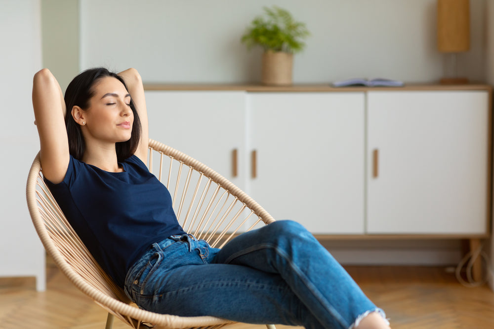 Woman relaxing in a woven chair at home with arms behind her head.