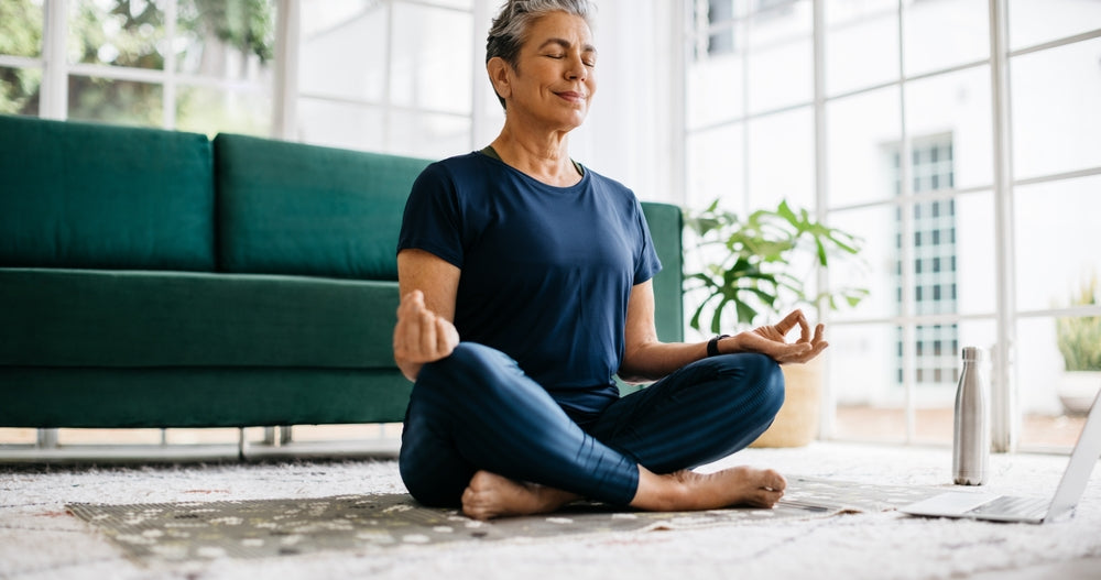 Calm woman in navy athletic wear sits cross-legged on a rug meditating indoors near a laptop and water bottle.