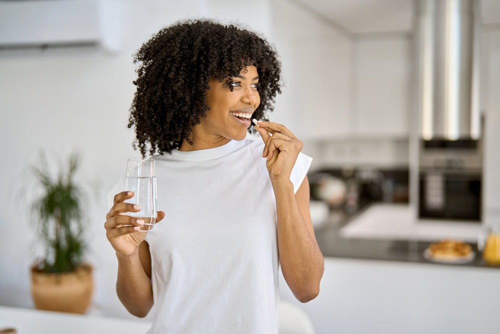 Smiling woman in a white shirt taking a pill with a glass of water in a bright kitchen.