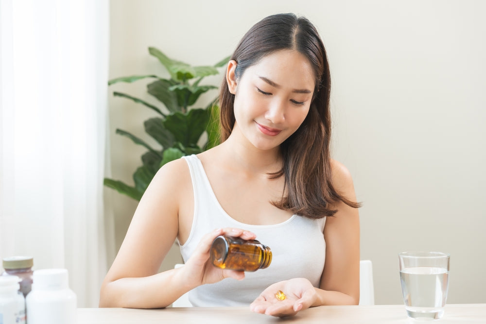 Young woman in a white tank top sits at a table, pouring a supplement capsule from a brown bottle into her hand beside a glass of water.