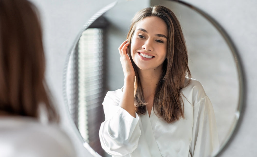 Smiling woman in a white robe gazes at her reflection in a round bathroom mirror.