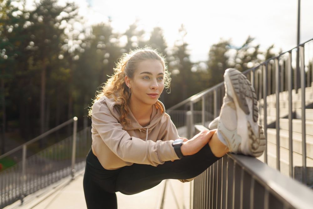 Woman in beige hoodie stretches her leg on a railing during an outdoor workout.