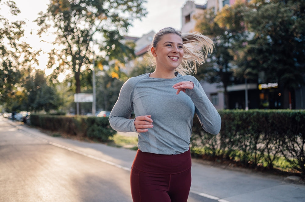 Smiling woman in a light gray long-sleeve top and burgundy leggings jogs along a sunlit city street.