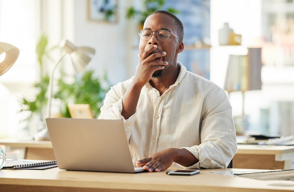 Man at a desk yawning beside a laptop in a bright office.