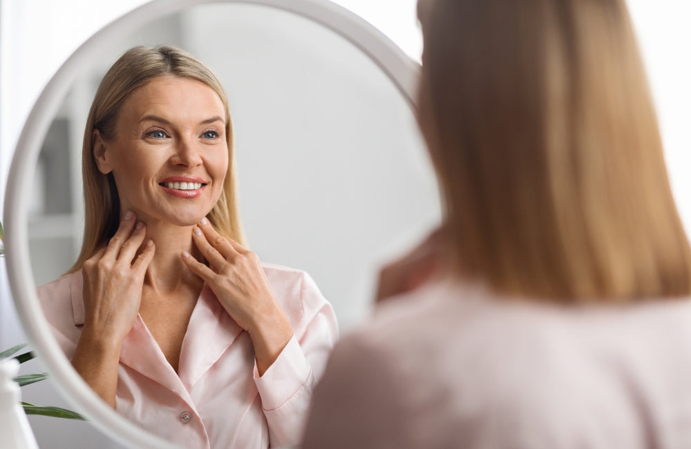 Smiling woman in a pink robe touches her neck while looking at her reflection in a round mirror for the Nutrition In Focus article.