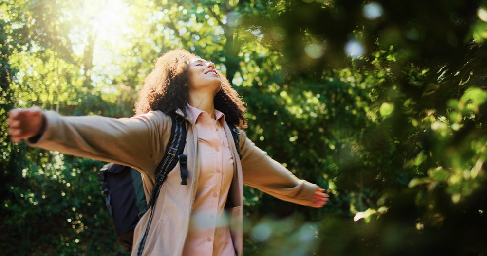 Woman with backpack arms outstretched in a sunlit forest setting.