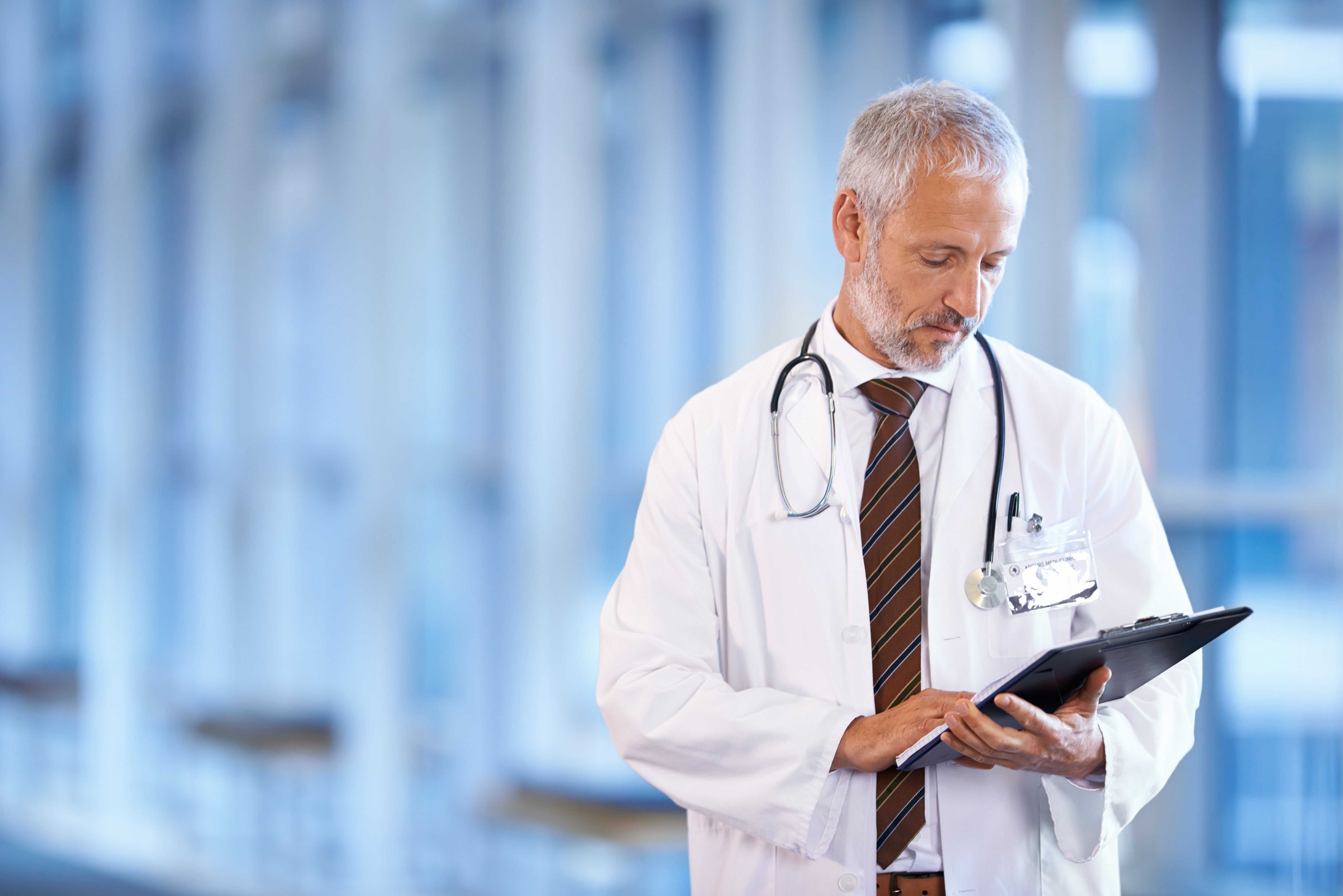 Practitioner in a white coat with a stethoscope, holding a tablet in a hospital setting.