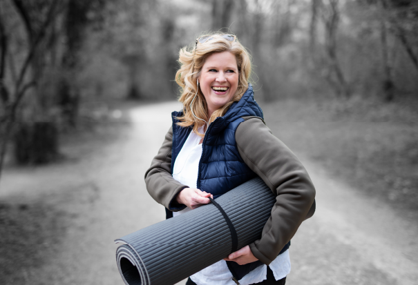 Smiling woman outdoors carrying a rolled yoga mat, dressed for exercise.