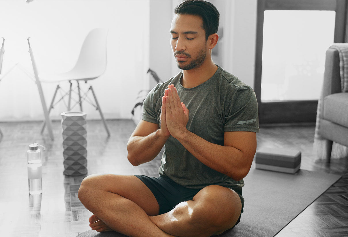 Man meditating cross-legged on a mat with hands in prayer position in a bright home.