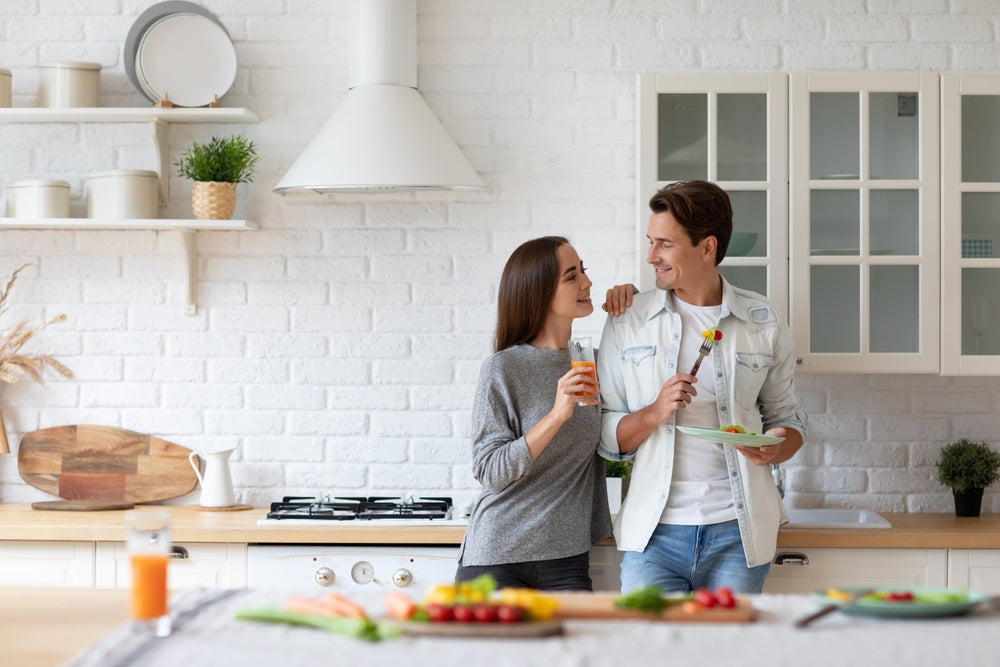 Couple in a bright white kitchen share a moment, one holds a glass while the other holds a plate with a veggie skewer.