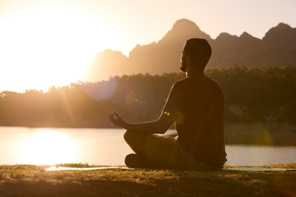 Man meditating on a mat beside a lake at sunset with mountains in the background.