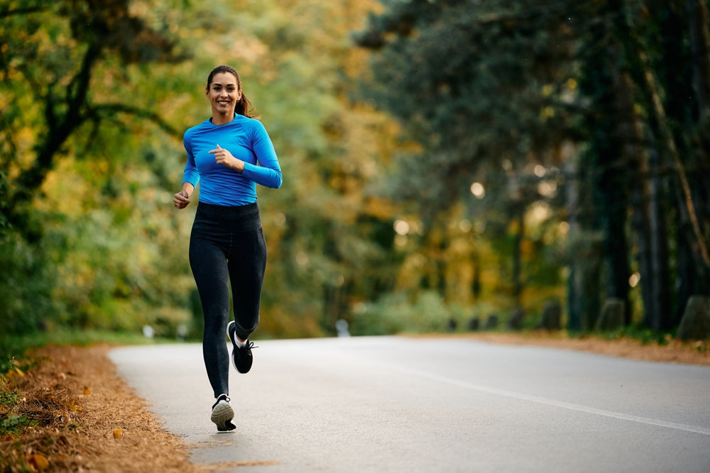 Woman running along a tree-lined autumn road in a blue long-sleeve top and black leggings.