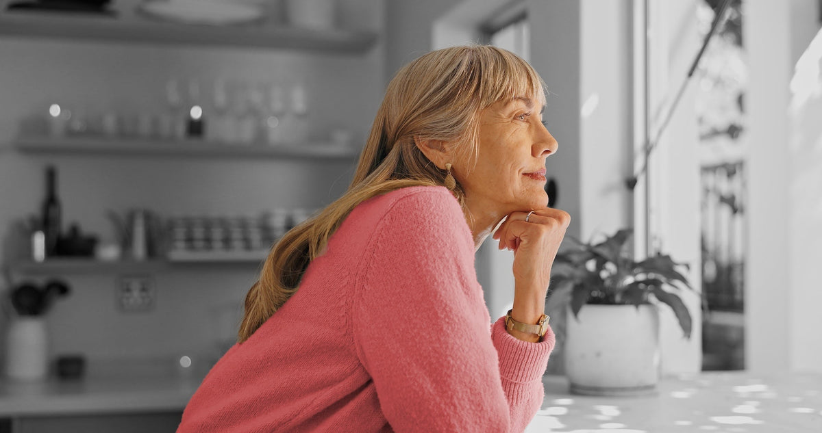 Older woman with blonde hair in a pink sweater rests her chin on her hand at a bright kitchen.