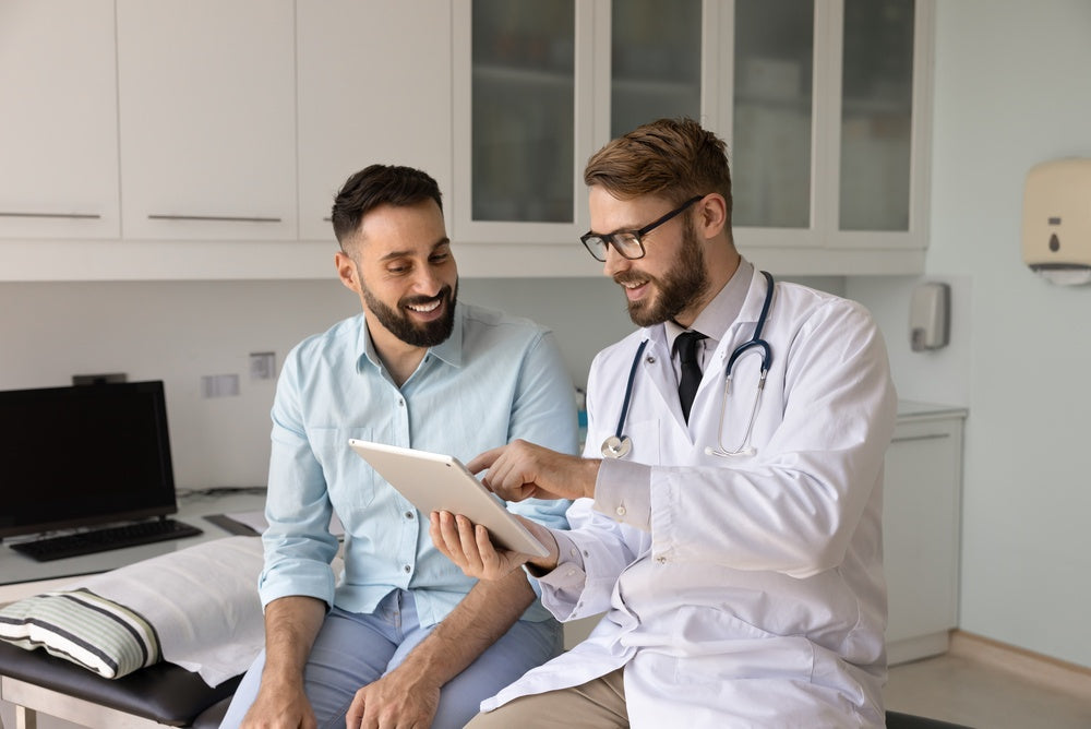 Doctor in white coat with stethoscope shows tablet to smiling male patient in a clinic.