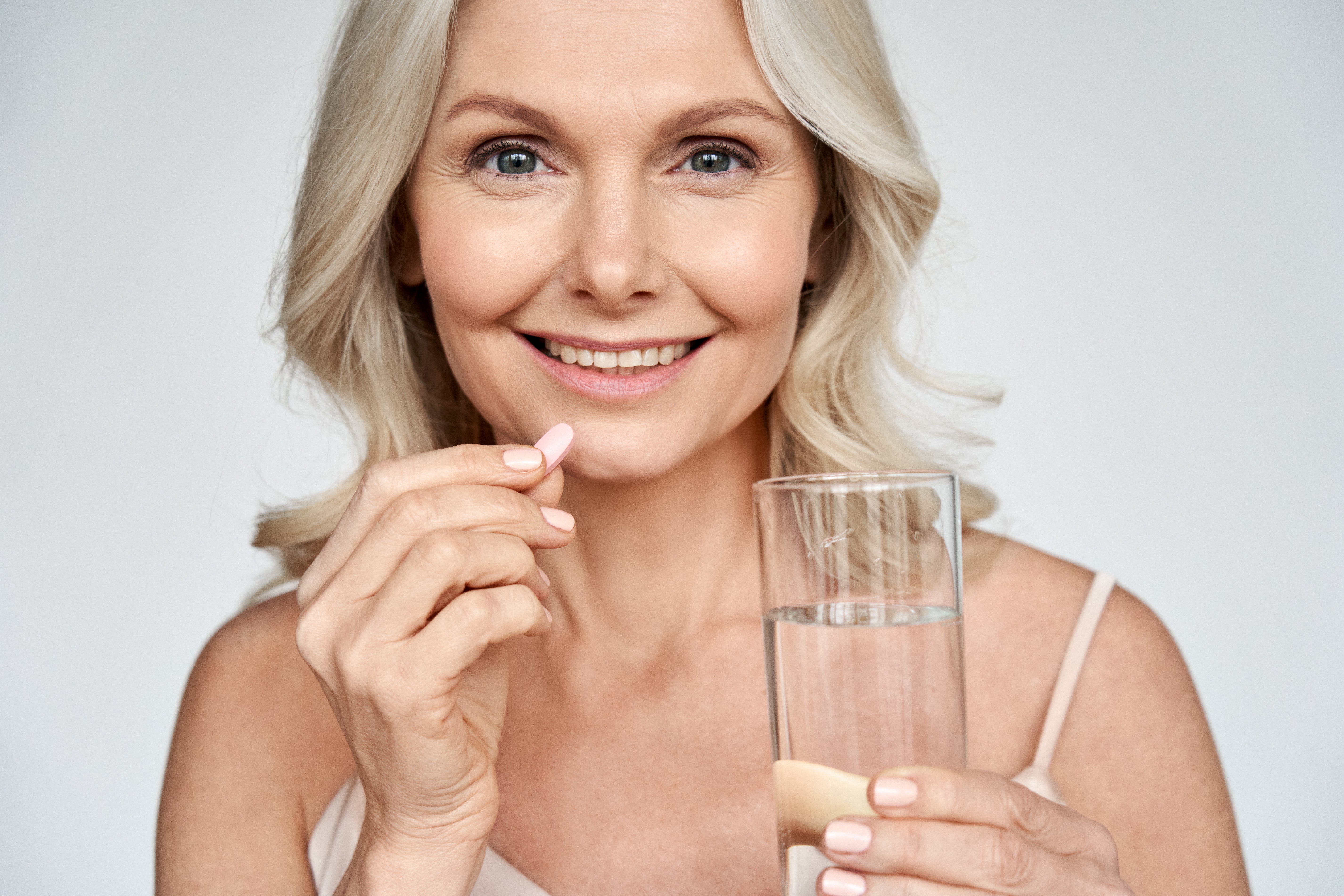 Woman holding a glass of water and a pill against a plain background